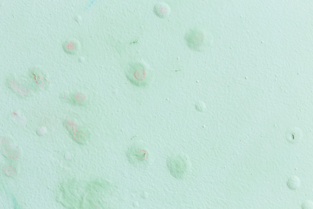 Parede verde-clara com textura irregular, pequenas bolhas, descascados e manchas suaves em tons de rosa e azul