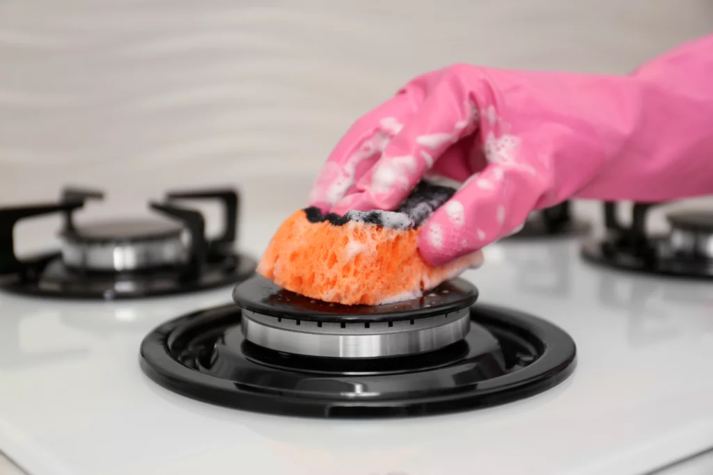 Mão com luva de borracha rosa limpa o queimador de um fogão com uma esponja ensaboada, removendo sujeira da superfície branca da cozinha.