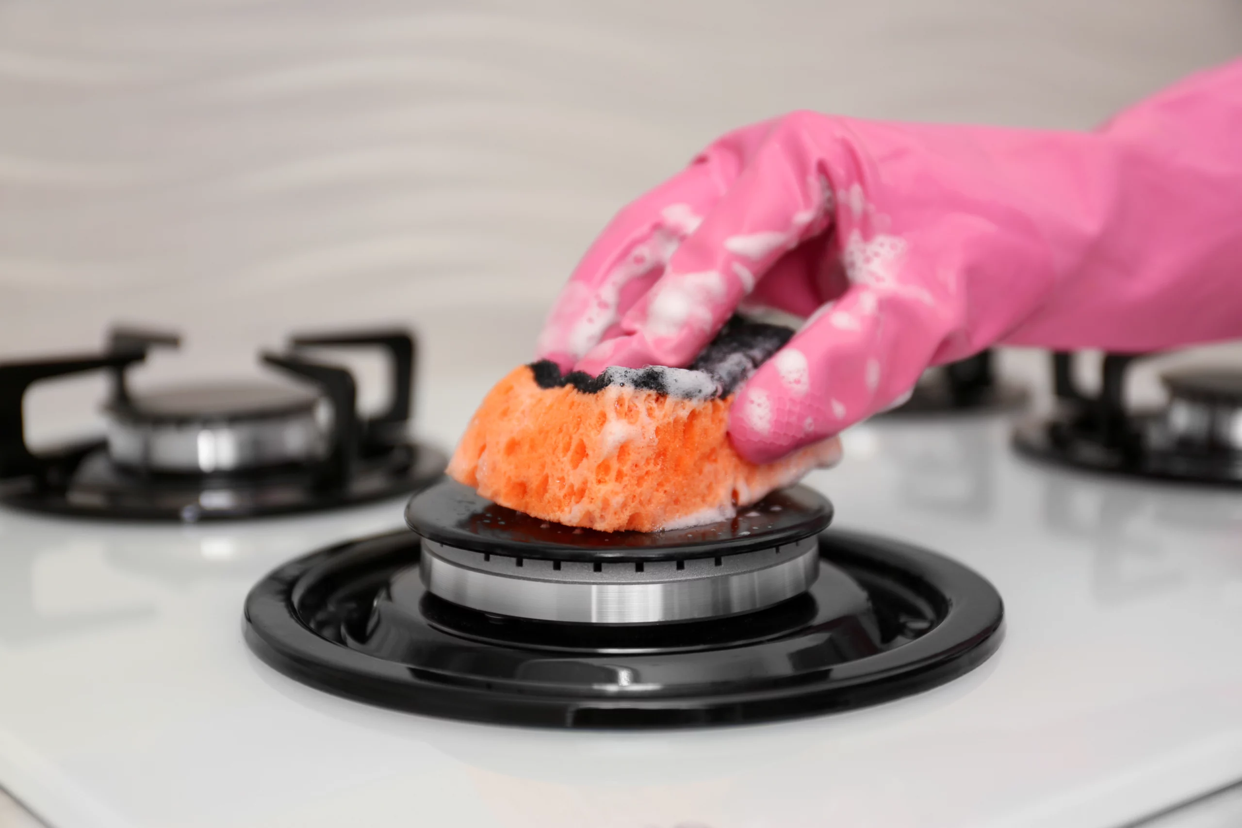 Mão com luva de borracha rosa limpa o queimador de um fogão com uma esponja ensaboada, removendo sujeira da superfície branca da cozinha.
