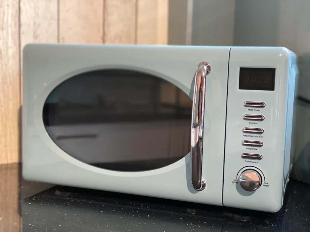 Micro-ondas azul-claro com porta oval e painel de controle, sobre bancada preta em uma cozinha.