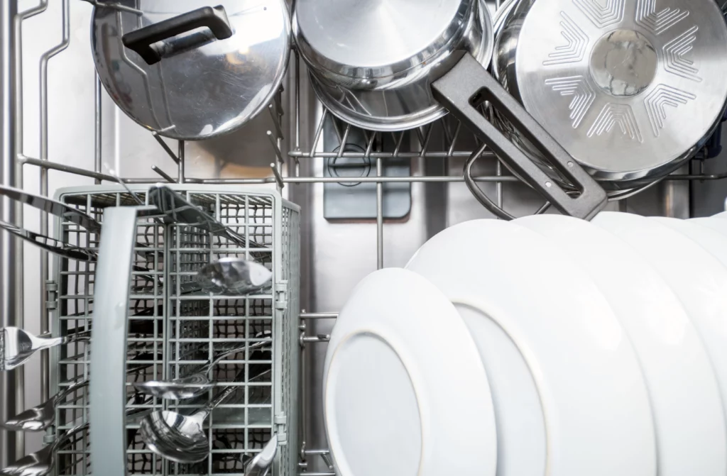 Interior de uma lava-louças aberta com panelas de inox, pratos brancos, talheres organizados no cesto e utensílios acomodados corretamente para o ciclo de lavagem.
