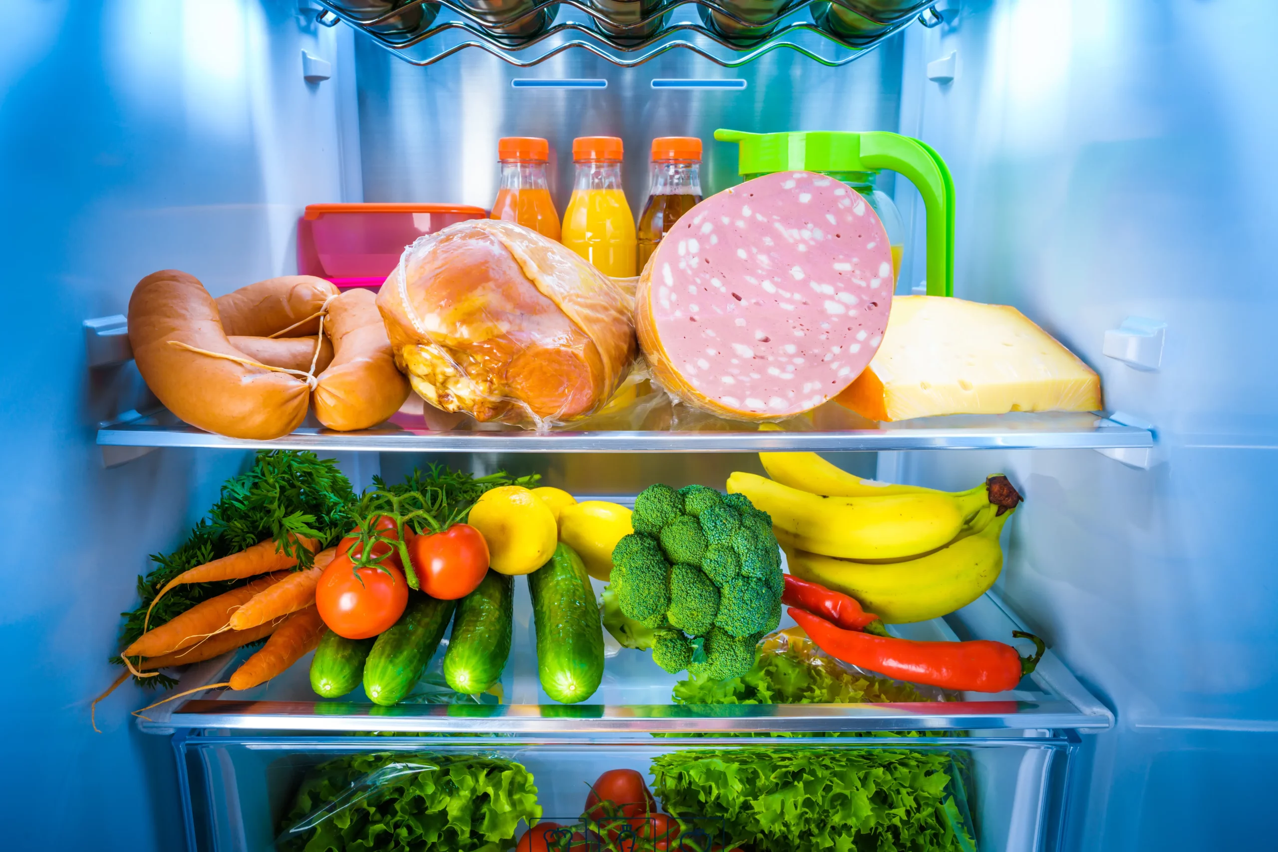 Interior de geladeira organizada com carnes, frios, queijo, bebidas, frutas, verduras e legumes frescos distribuídos nas prateleiras.