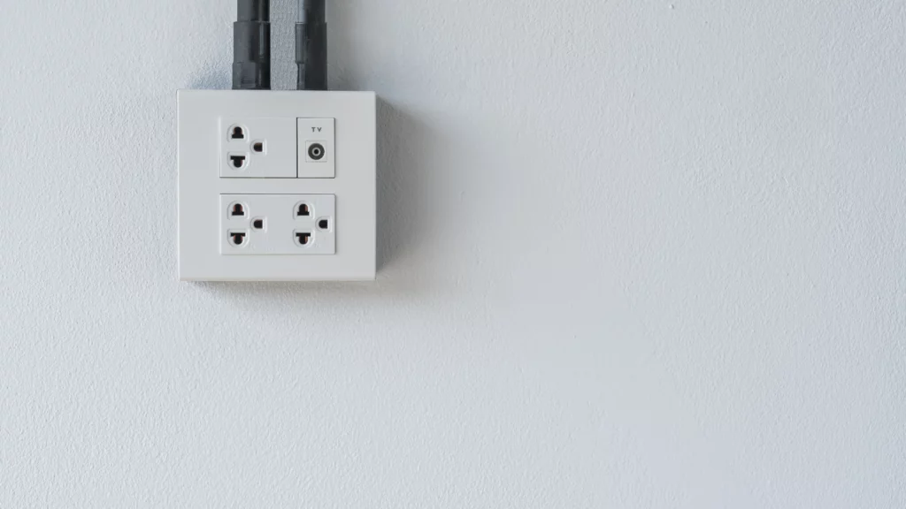 Tomada elétrica branca com entradas para plugues e TV, instalada em uma parede branca, com dois cabos pretos conectados na parte superior.