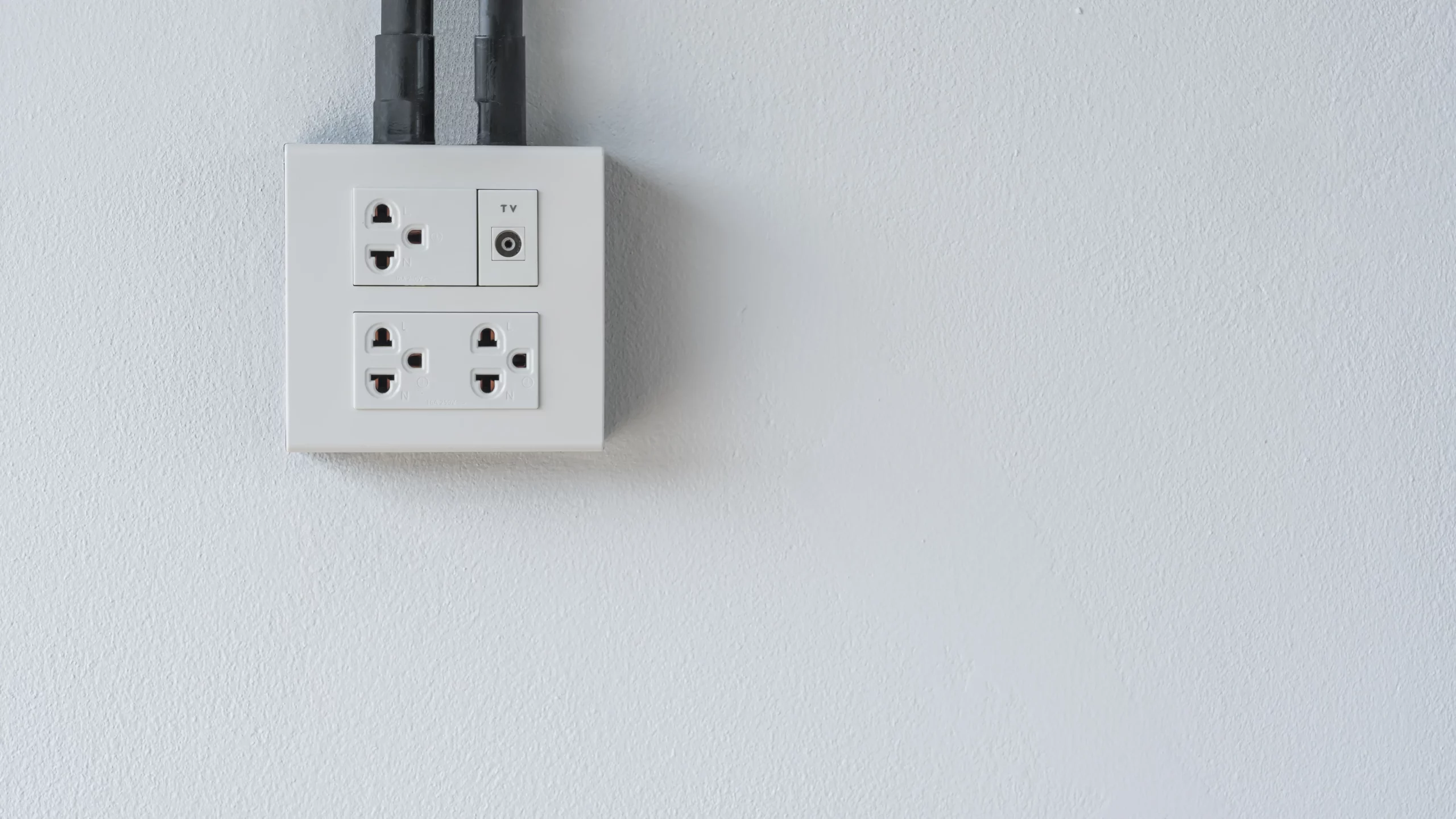 Tomada elétrica branca com entradas para plugues e TV, instalada em uma parede branca, com dois cabos pretos conectados na parte superior.